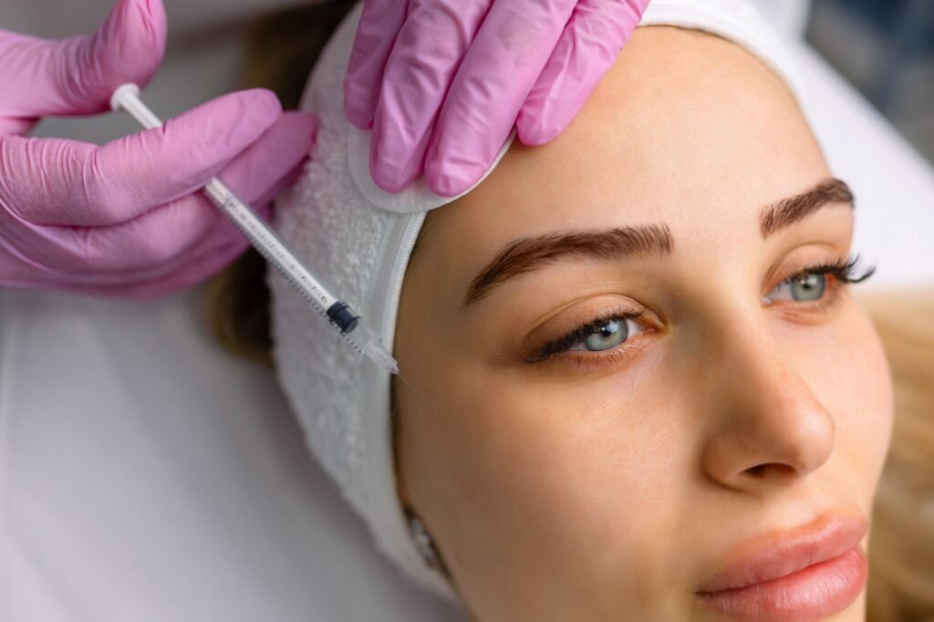 Close up of hands of young cosmetologist injecting botox in female face. She is standing and smiling